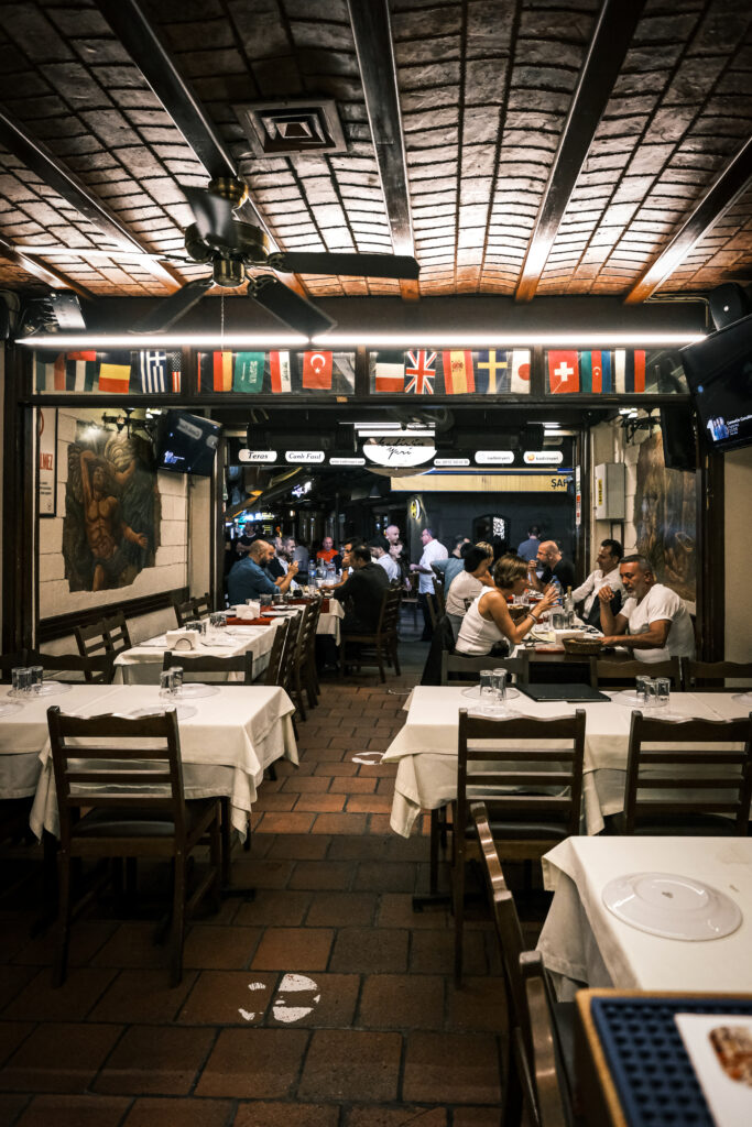 Loş ve sıcak ışıkta çekilmiş tarihi meyhane masası, üzerinde Beyoğlu En İyi Meyhane Deneyimini simgeleyen kadeh, Topik ve Lakerda mezeleri.